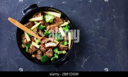 Roasted pork chunks mixed with vegetables: broccoli and corn cobs on a black baking dish on a dark concrete background view from above, horizontal ori Stock Photo