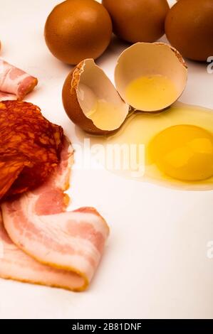 Broken chicken egg and scattered eggs, slices of sliced sausage and bacon on a white background. Close up Stock Photo