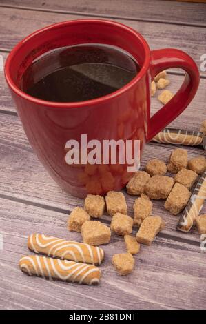 Red ceramic tea mug, cookies and pieces of cane sugar on a wooden background. Close up Stock Photo