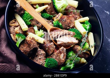 Leftover steak kebabs with vegetables on a black baking dish: broccoli, corn, onion, and garlic on a dark concrete background, close-up, horizontal or Stock Photo