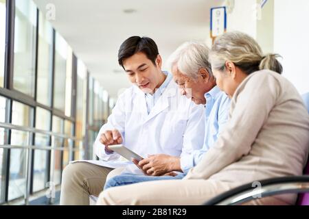 young asian doctor discussing test result and diagnosis with senior couple patients using digital tablet in hospital hallway Stock Photo