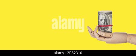 Wooden hand with money isolated on yellow background. Roll of one hundred dollar bills. 100 USD banknotes, American Dollars currency. Long wide banner Stock Photo