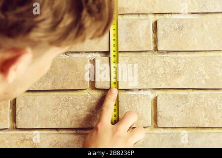Construction and measurement concept. Man holds yellow self retracting tape measure. Guy measures brick wall with builders ruler. Worker with roulette on beige brick background, defocused Stock Photo