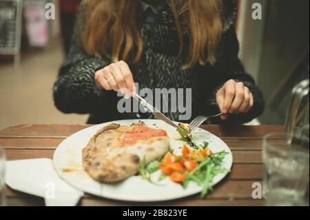 A young woman wearing a winter coat is eating a calzone pizza at a table outdoors in an arcade Stock Photo