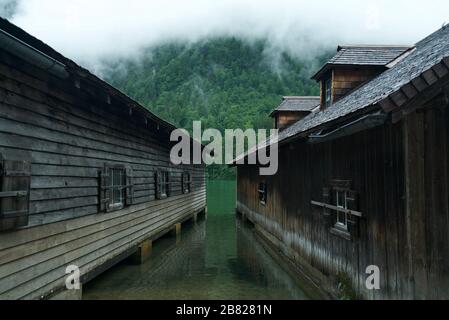 Boathouse / Boathouses by the water surrounded by mountains and clouds at Lake Königssee (Berchtesgaden National Park). Wooden cabins by the lake. Stock Photo