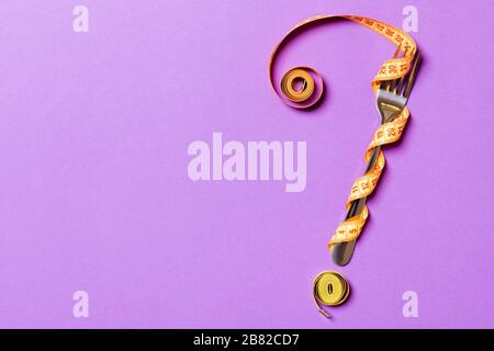 Top view of fork and measure tape in form of question mark on purple background. Overeating concept and copy space. Stock Photo