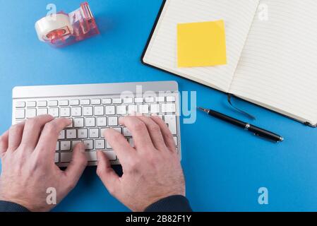 top view of person typing on computer keyboard against blue desk background with open notebook Stock Photo