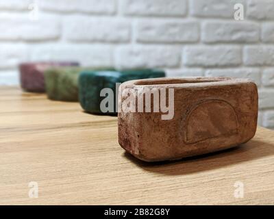 Brown concrete pot on wooden table with emarald, green and purple pots behind and white brick wall on the background Stock Photo