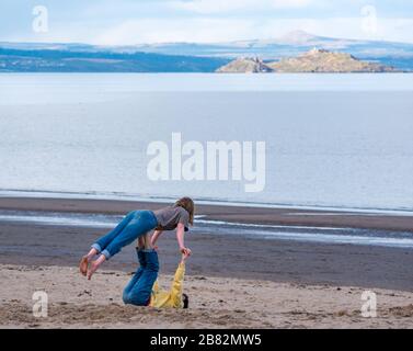 Portobello beach, Edinburgh, Scotland, United Kingdom. 19th Mar, 2020. Social distancing, or not? On a sunny Spring day, people are out enjoying the seaside. A young couple practise acrobatics on the beach on the shore of the Firth of Forth Stock Photo