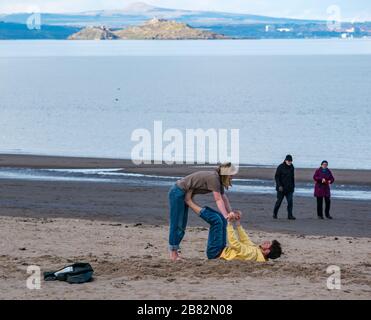 Portobello beach, Edinburgh, Scotland, United Kingdom. 19th March 2020. On a sunny Spring day, people are out enjoying the seaside. A young couple practise acrobatics on the beach on the shore of the Firth of Forth during the Covid-19 pandemic Stock Photo