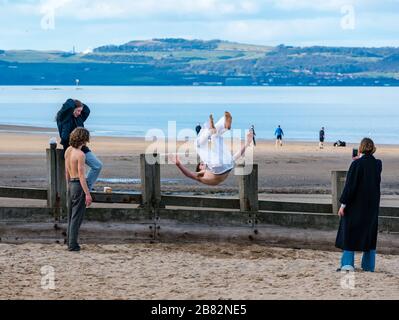 Portobello beach, Edinburgh, Scotland, United Kingdom. 19th March 2020. On a sunny Spring day, people are out enjoying the seaside.. A young man does a flip while his friends look on during the Covid-19 pandemic Stock Photo