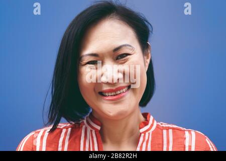 Senior Asian woman portrait against blue background - Smiling Chinese female having fun posing in front of camera - Mature people lifestyle concept Stock Photo