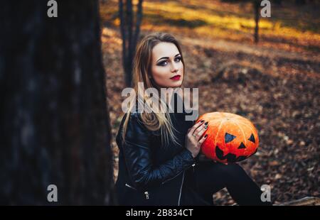 beautiful young blond woman with extravagant make-up in a black leather jacket with wide open eyes and an open mouth with a pumpkin in hands Stock Photo