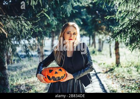 beautiful young blond woman with extravagant make-up in a black leather jacket with wide open eyes and an open mouth with a pumpkin in hands Stock Photo
