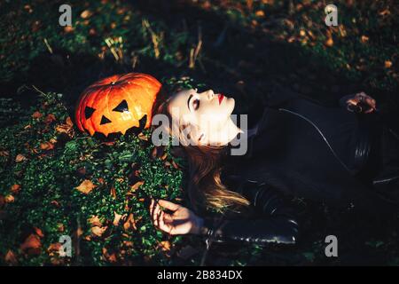 beautiful young blond woman with extravagant make-up in a black leather jacket with wide open eyes and an open mouth with a pumpkin in hands Stock Photo