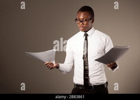 Young positive handsome african businessman in stylish shirt, tie and glasses standing with documents over grey background. Trendy business look and o Stock Photo