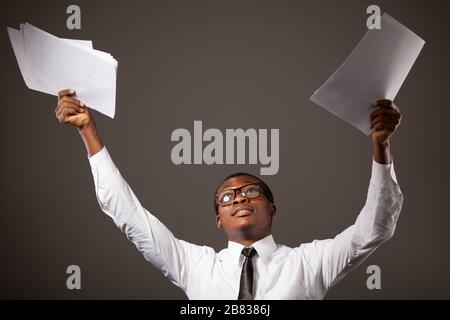 Young positive handsome african businessman in white shirt, tie and glasses standing with documents in raised hands over grey background. Trendy busin Stock Photo