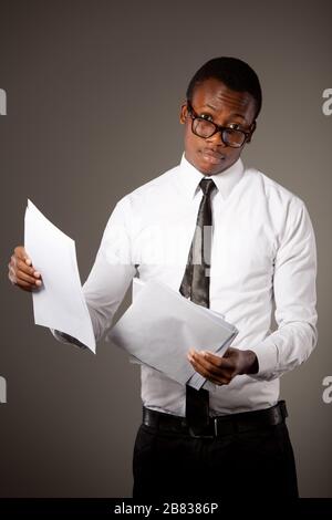 Young positive handsome african businessman in stylish shirt, tie and glasses standing with documents over grey background. Trendy business look and o Stock Photo