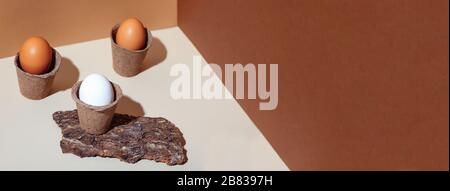 Three multicolored eggs in pots. Natural materials: bark of wood. Geometric lines, brown background. Banner Stock Photo