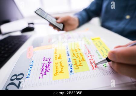 Close-up Of A Businessman Holding Cellphone Writing Schedule In The Diary With Pen Stock Photo
