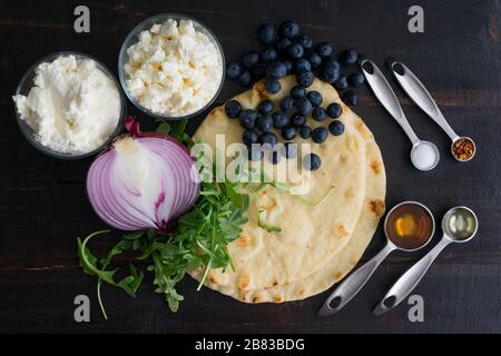 Ingredients for Blueberry, Feta and Honey-Caramelized Onion Naan Pizza: Naan flatbreads, arugula, and other ingredients arranged on a wood table Stock Photo