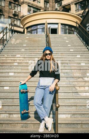 Stylishly dressed woman in blue denim jumpsuit posing with skateboard. Street photo. Portrait of girl holding skateboard. Lifestyle, youth concept Stock Photo