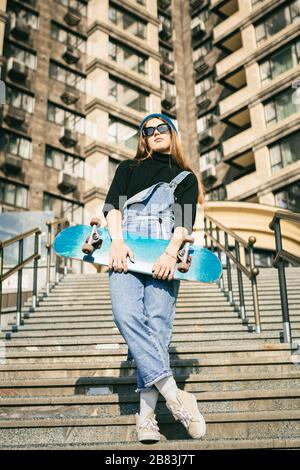 Young caucasian woman posing on street with skateboard in hands. Teenager girl in blue jeans extreme sports in an urban environment. Theme of youth Stock Photo