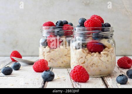 Overnight oats with fresh blueberries and raspberries in jars on a rustic white wood background Stock Photo
