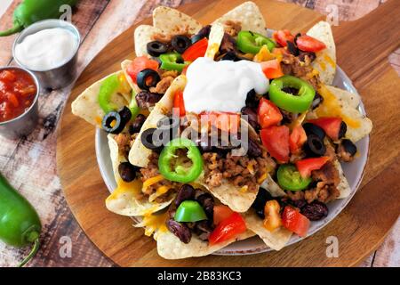 Mexican nacho chips topped with sour cream, ground meat, jalapenos, tomatoes, beans and melted cheese on a wooden paddle board Stock Photo