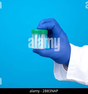 doctor in a white coat and blue sterile latex gloves holds a plastic jar for stool analysis, blue background Stock Photo