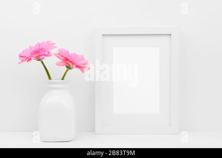 Mock up white frame and pink daisy flowers in vase on a shelf or desk. White color scheme. Portrait frame orientation. Stock Photo