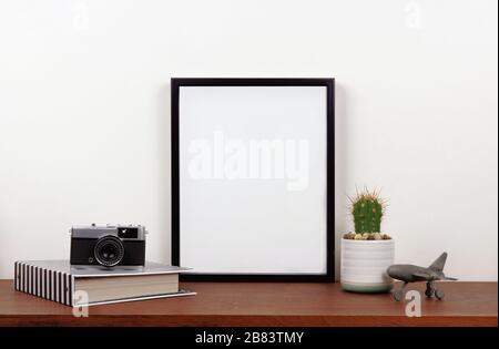 Mock up black frame with travel theme decor on a shelf or desk. Wood shelf and white wall. Portrait frame orientation. Stock Photo