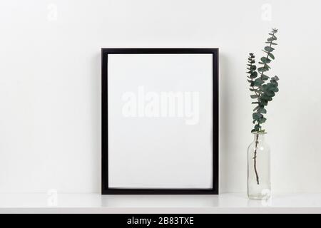 Mock up black frame with vase of branches on a shelf or desk. White shelf and wall. Portrait frame orientation. Stock Photo