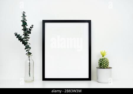 Mock up black frame with cactus and branches on a shelf or desk. White shelf and wall. Portrait frame orientation. Stock Photo