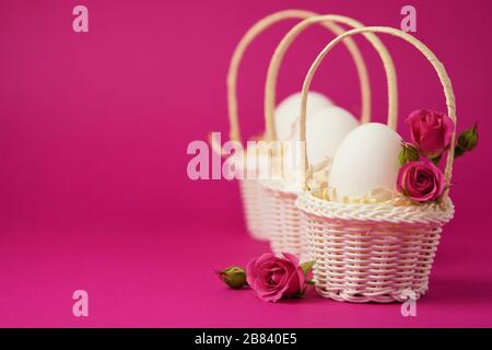 White chicken eggs in a white wicker basket covered with straw and small pink roses on a bright pink background. Holiday card with space for text Stock Photo