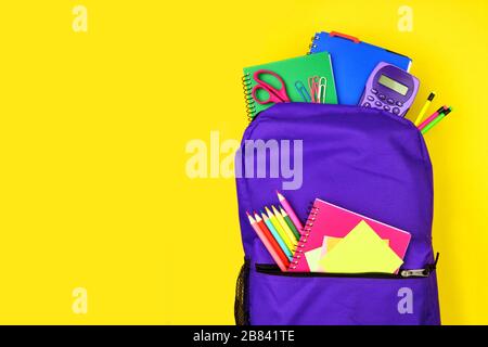 Purple backpack full of school supplies against a bright yellow background. Close up, top view with copy space. Stock Photo
