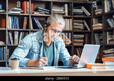Focused man student wear headset study online make notes in library. Stock Photo