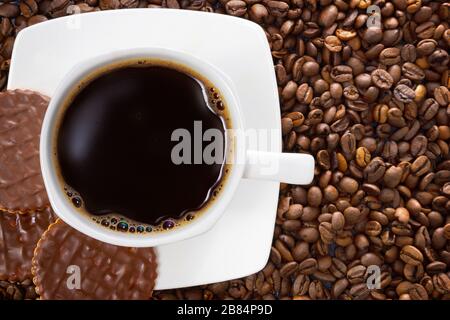 White cup of hot coffee on a background of brown beans. Three chocolate biscuits. Stock Photo