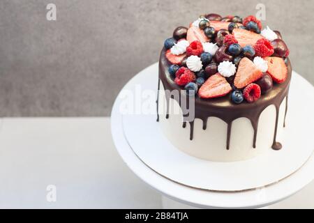 White cake with melted dark chocolate, fresh strawberries, blueberries, raspberries and cherries on gray background Stock Photo