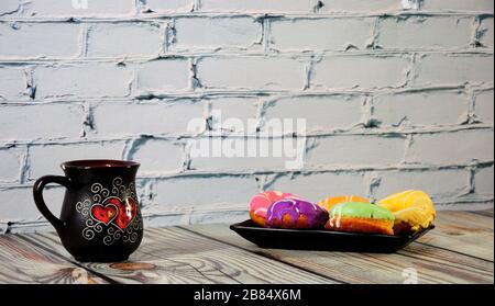 A rectangular plate with five multi-colored donuts and a black ceramic coffee mug stand on a wooden table. Close-up. Stock Photo