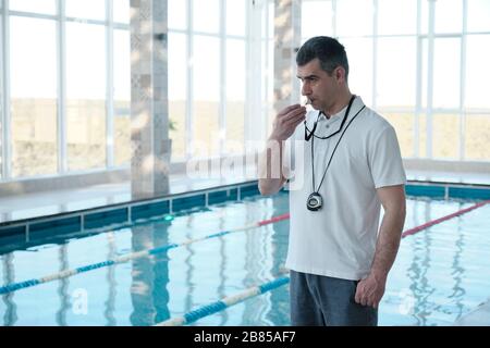 Experienced gray-haired male coach with stopwatch hanging on neck blowing whistle to swimmer in indoor pool Stock Photo