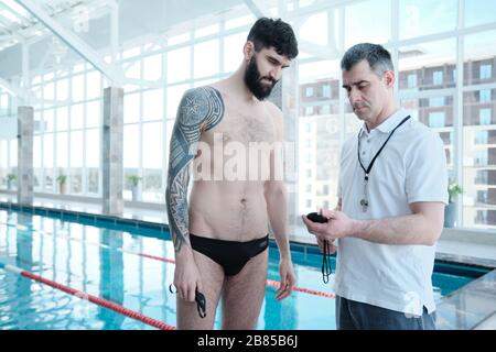 Bearded man with tattoo standing at pool and talking about lap time to swimming coach Stock Photo