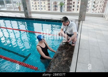 Mature coach with whistle crouching on poolside and giving advice to swimmer how to improve lap time
