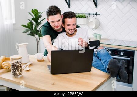 Happy gay male couple embracing while using laptop, having a good time together in kitchen at home. Happy gay couple concept. Stock Photo