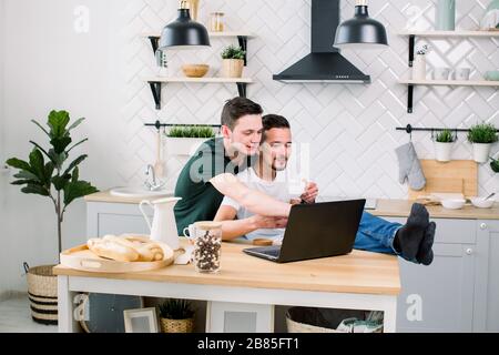 Happy gay male couple embracing while using laptop, having a good time together in kitchen at home. Happy gay couple concept. Stock Photo