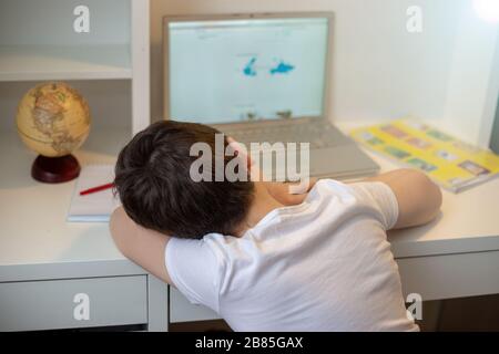 A schoolboy in a white T-shirt fell asleep behind a monitor from a laptop, holding his head in his hands. Home lessons. Exam preparation. hard to lear Stock Photo