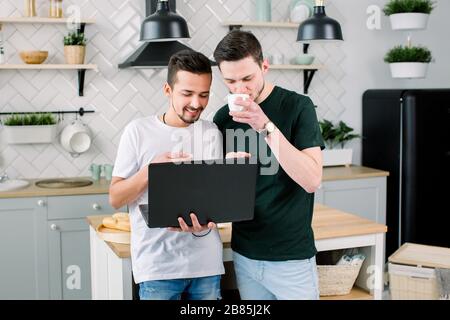 Two happy friends having fun while using laptop together. Happy smiling gay couple using laptop at the kitchen at home. Stock Photo
