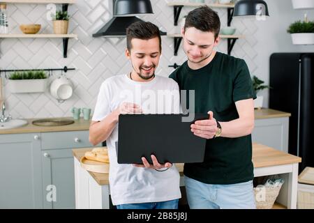 Two happy friends having fun while using laptop together. Happy smiling gay couple using laptop at the kitchen at home. Stock Photo