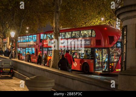 Side view, red double decker London buses parked, Trafalgar Square, central London UK at night. Bus passengers getting on and off public bus transport. Stock Photo