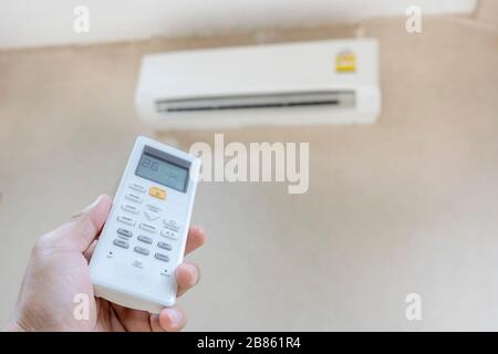 The hand that is holding the remote to control the air conditioner requires a cooler temperature. The background is an air conditioner that is running Stock Photo